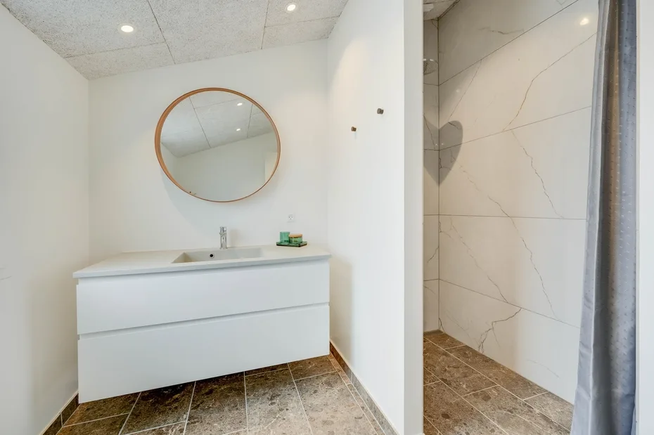 Un baño luminoso con tocador blanco, espejo redondo y ducha alicatada.