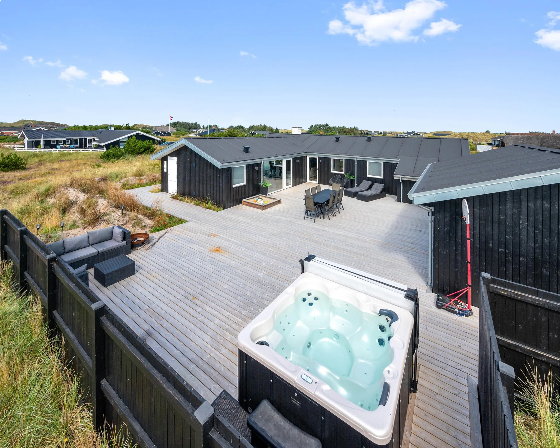 Una casa moderna se abre a una amplia terraza de madera con asientos al aire libre y un jacuzzi.