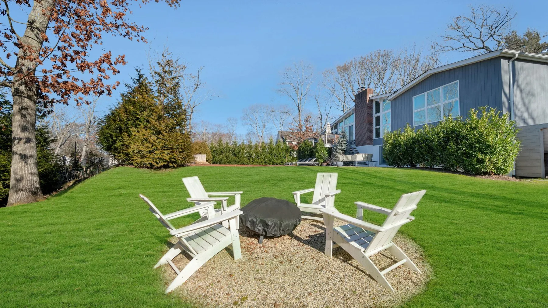 Un patio con césped tiene asientos al aire libre alrededor de un fogón junto a una casa.