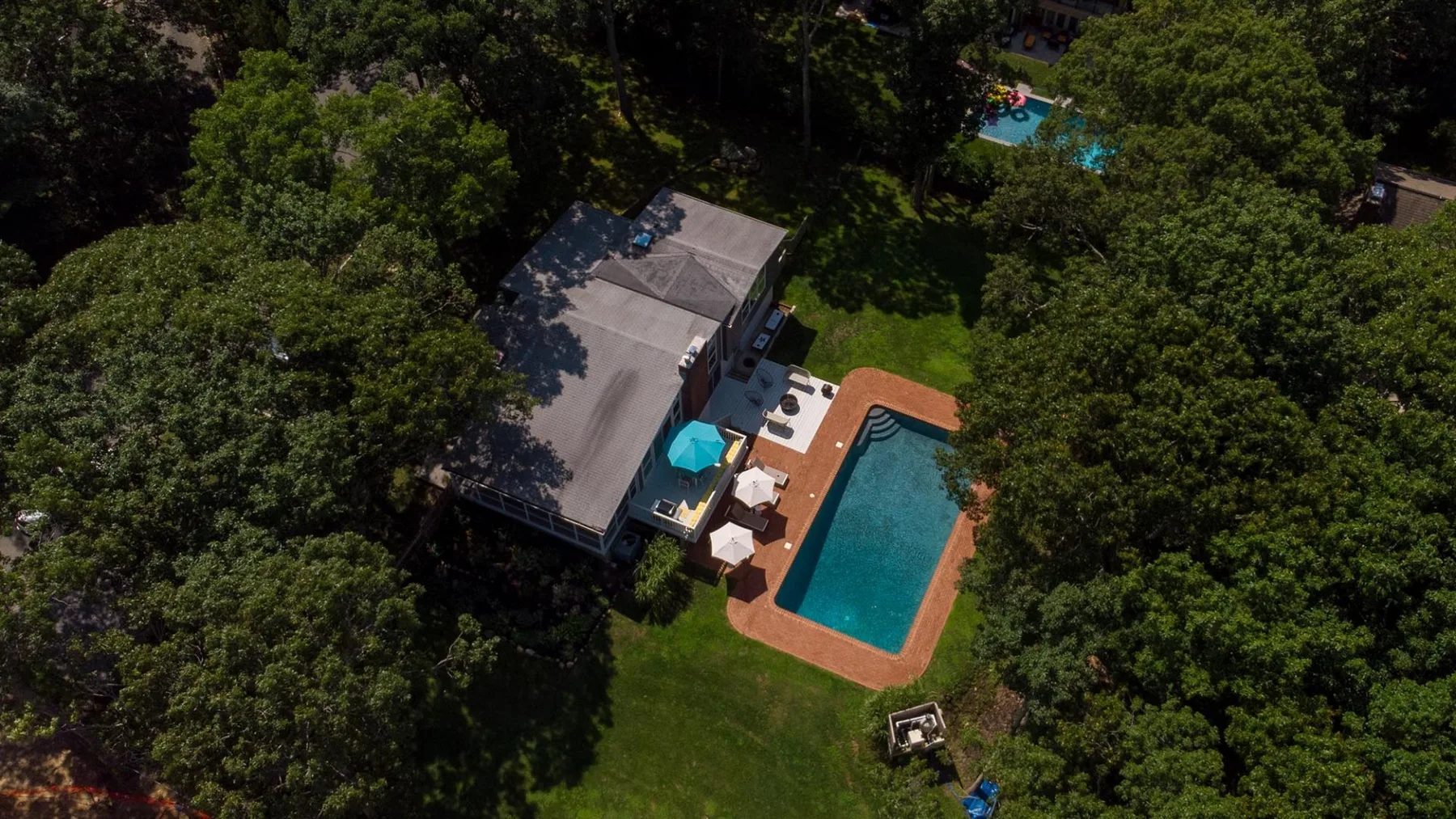 An aerial view shows a house beside a swimming pool amid dense trees.