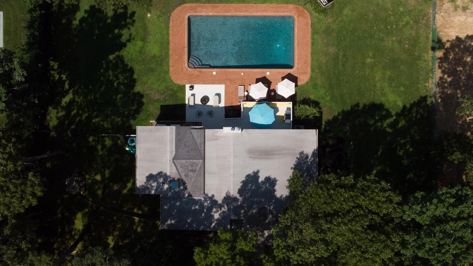 An aerial view shows a swimming pool beside a lawn and paved patio.