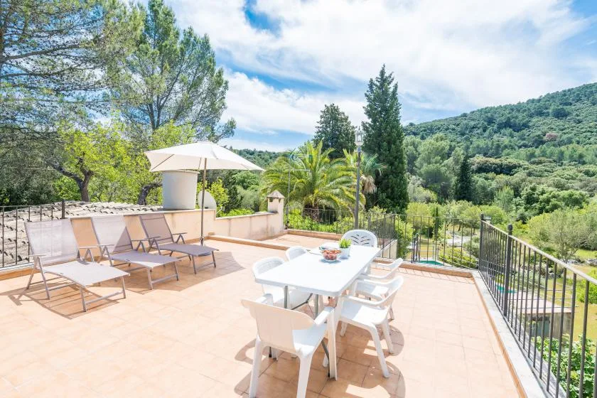 A sunlit outdoor terrace has seating, a dining table, and views of green hills.