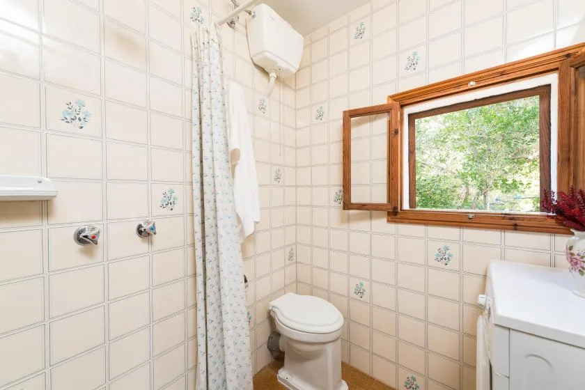 A tiled bathroom with a toilet, shower curtain, and window overlooking greenery.