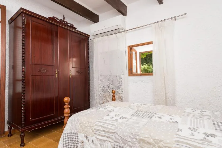 A bedroom with a large wooden wardrobe and a bed beside a small window.