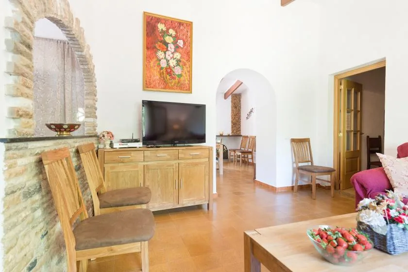 A bright living room has wooden seating, a television, and a dining area beyond an archway.