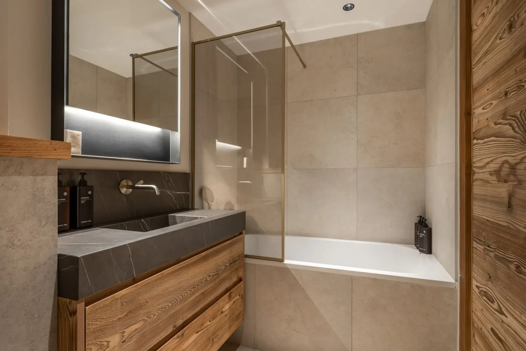 Baño moderno con un mueble de lavabo de madera, encimera gris, un espejo grande y una bañera con mampara de vidrio y azulejos beige.