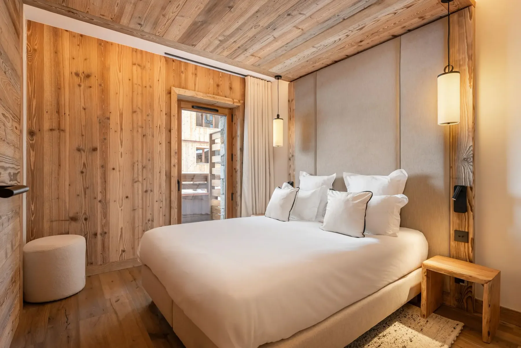 Dormitorio acogedor con paredes y techo de madera, una mullida cama blanca, lámparas colgantes y un pequeño taburete de madera junto a la cama. Luz natural procedente de una ventana.