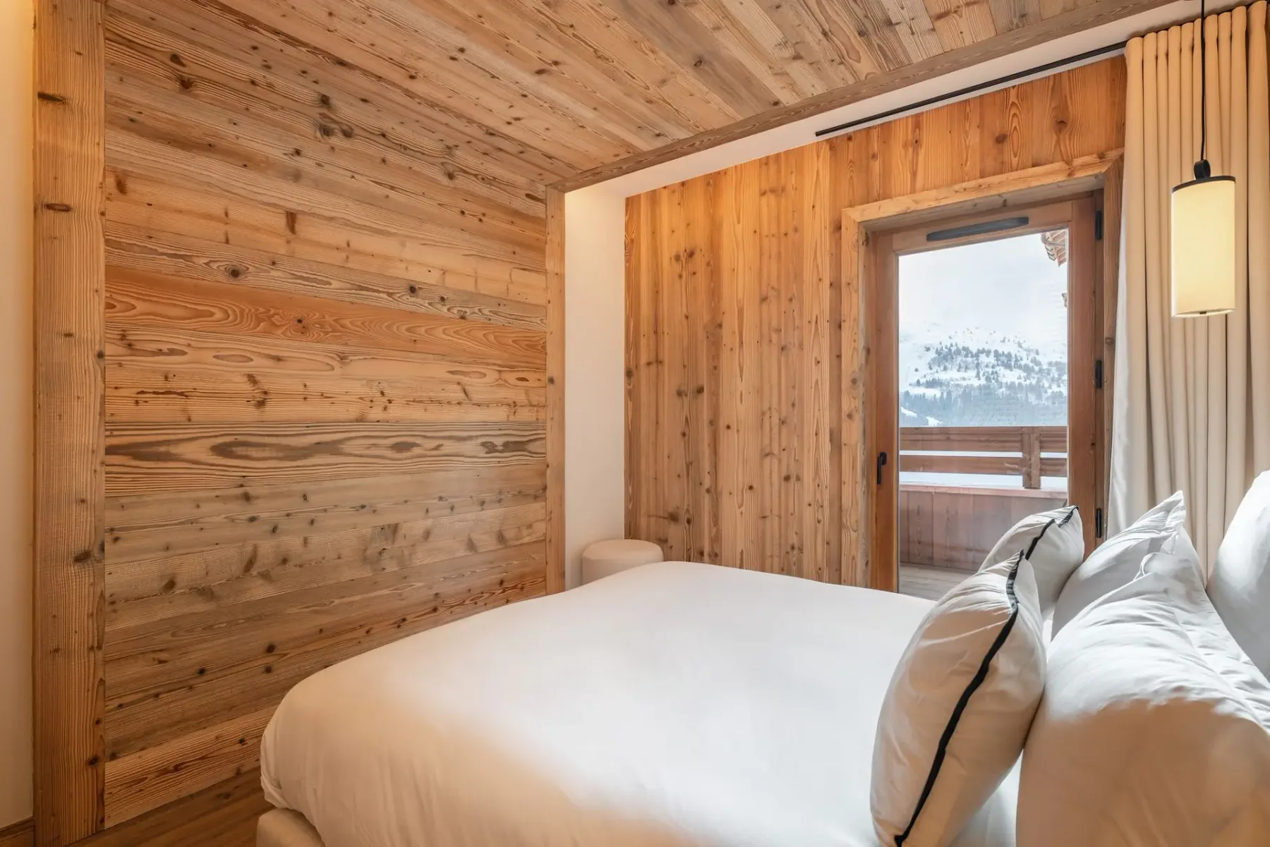 Dormitorio acogedor con paredes y techo de madera, una mullida cama blanca, lámparas colgantes y un pequeño taburete de madera junto a la cama. Luz natural procedente de una ventana.