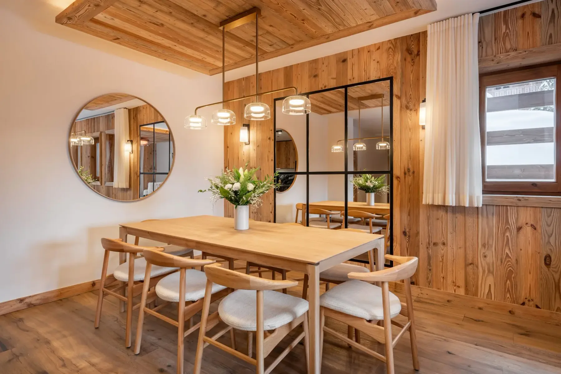 Comedor de madera con una mesa grande, sillas, lámparas colgantes y un espejo redondo en la pared.