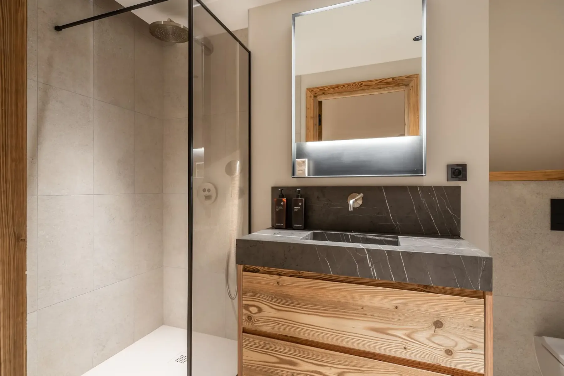 Baño moderno con una ducha con mampara de vidrio, encimera de piedra, muebles de madera y un espejo retroiluminado en una pared de color crema.