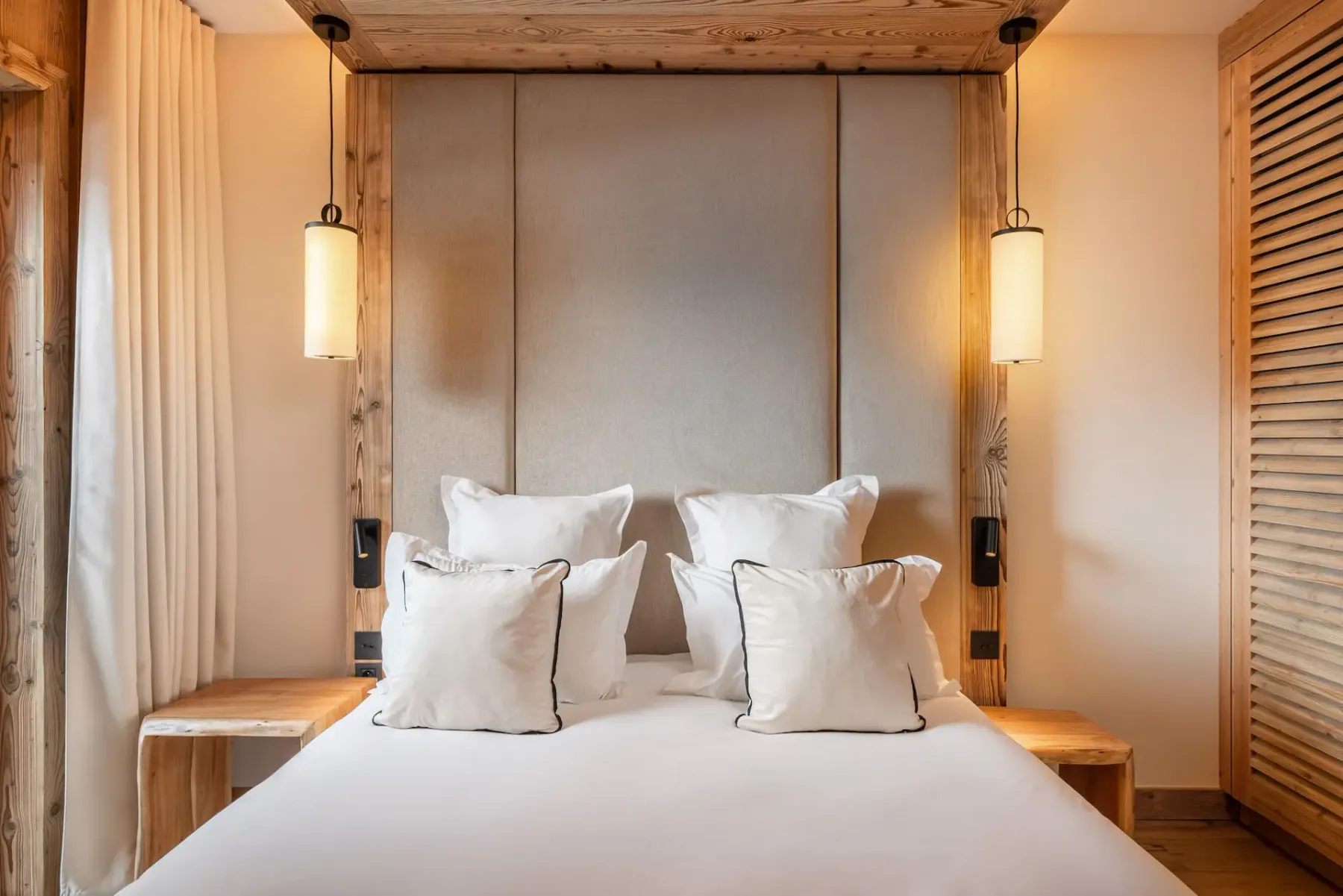 Dormitorio acogedor con paredes y techo de madera, una mullida cama blanca, lámparas colgantes y un pequeño taburete de madera junto a la cama. Luz natural procedente de una ventana.