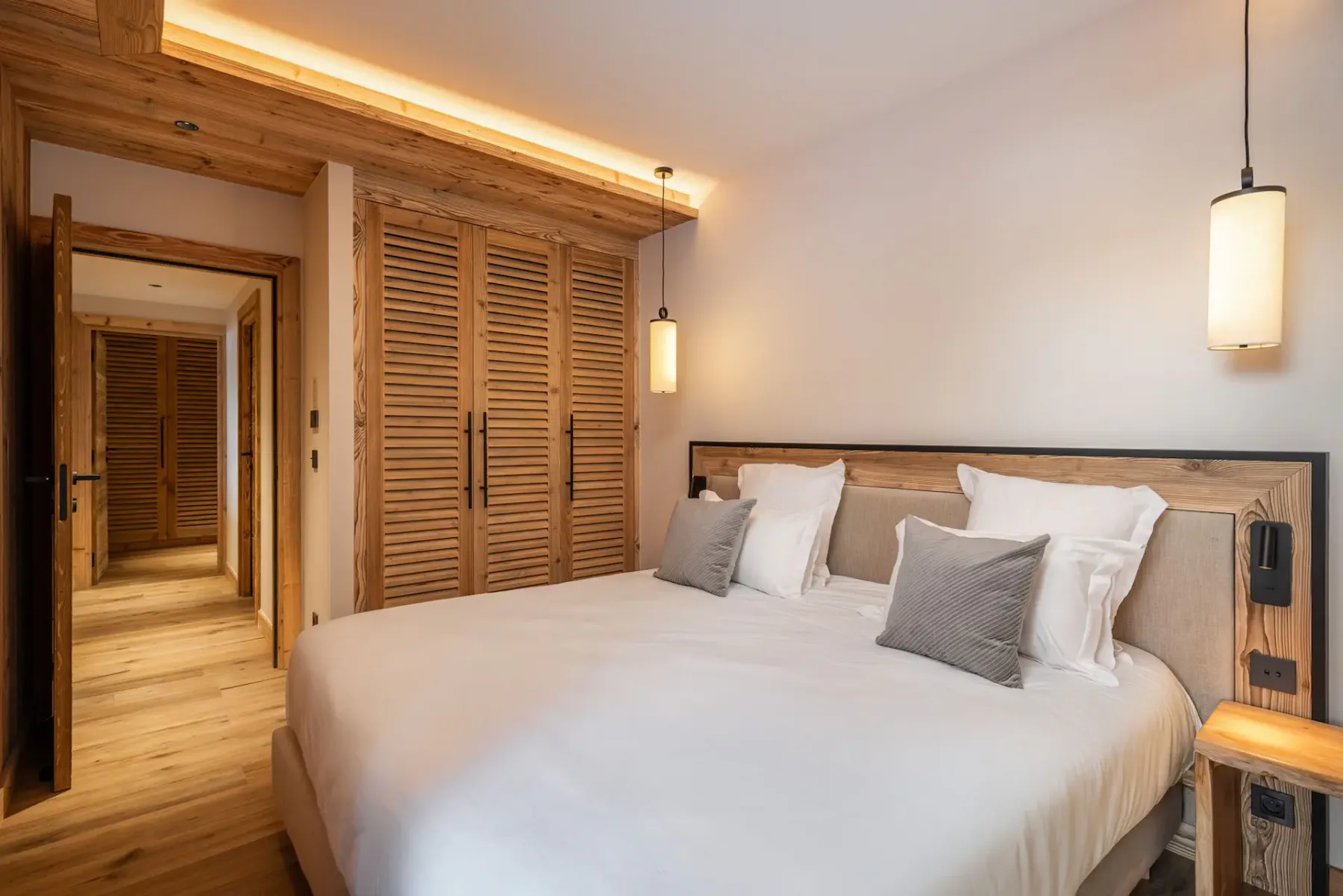 Dormitorio moderno con una mullida cama doble, detalles de madera, iluminación tenue y colores neutros que crean un ambiente cálido y acogedor.