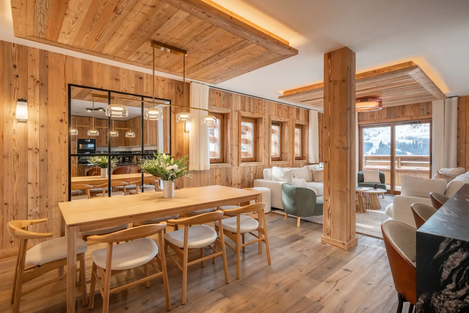Cálida zona de estar y comedor con predominio de la madera, una gran mesa de comedor, asientos acogedores y vistas panorámicas de un paisaje nevado a través de las ventanas.