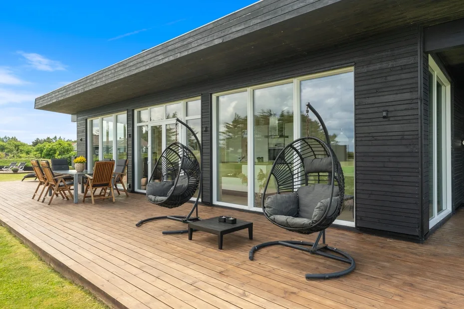 Una casa moderna se abre a una terraza de madera con sillas colgantes y asientos al aire libre.