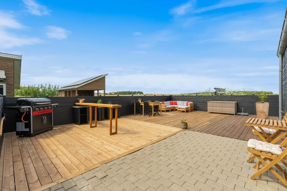 Una terraza exterior tiene suelo de madera, asientos y una parrilla.