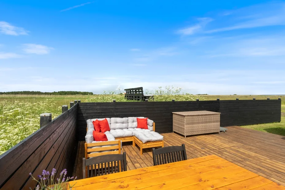 Una terraza de madera tiene asientos acolchados y una mesa junto a un campo abierto.