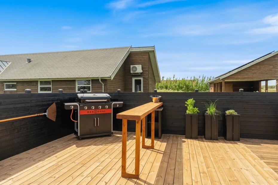 Una terraza de madera cuenta con una parrilla exterior, una mesa estrecha y jardineras.