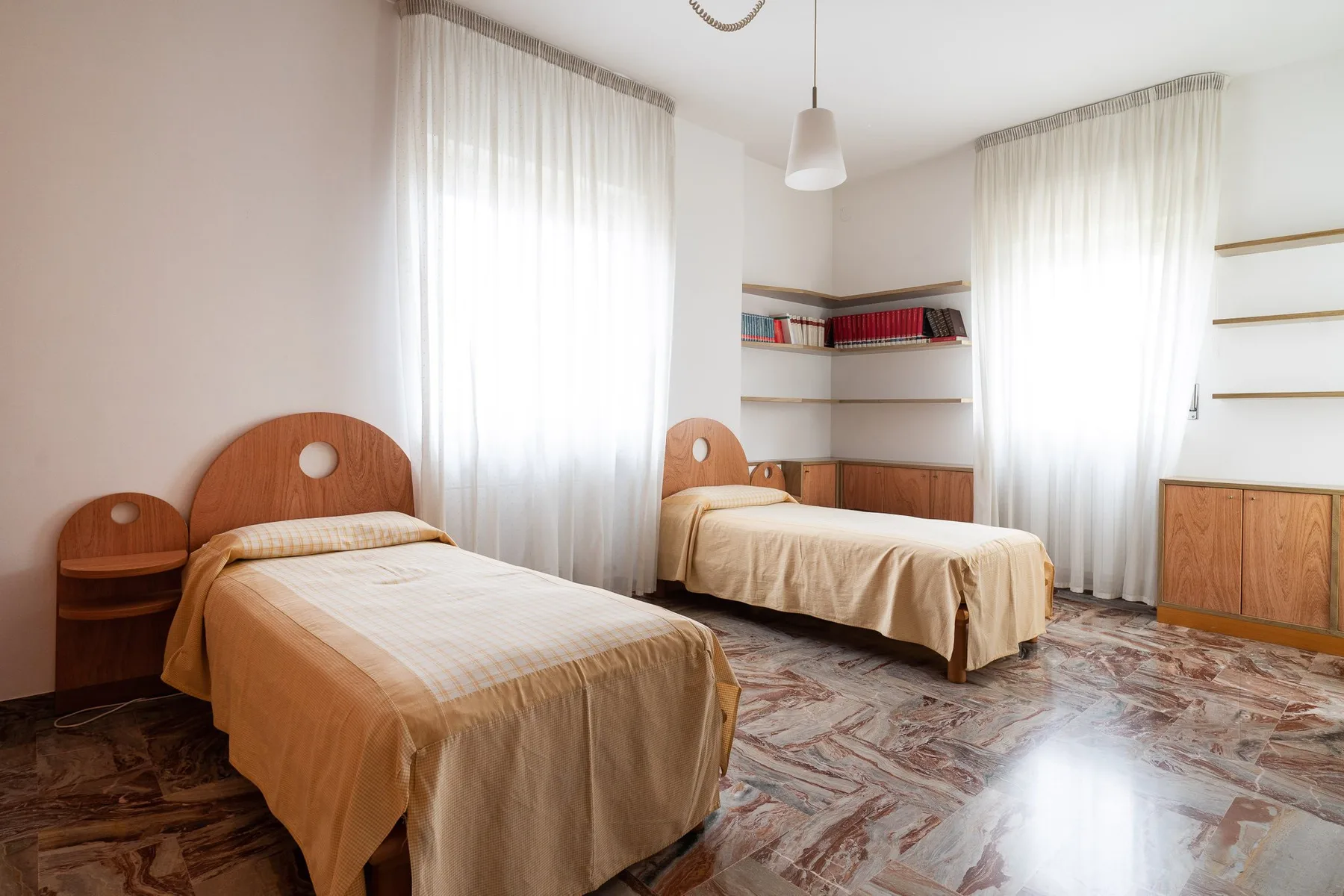Un dormitorio con dos camas individuales, ventanas altas con cortinas y estantes empotrados.