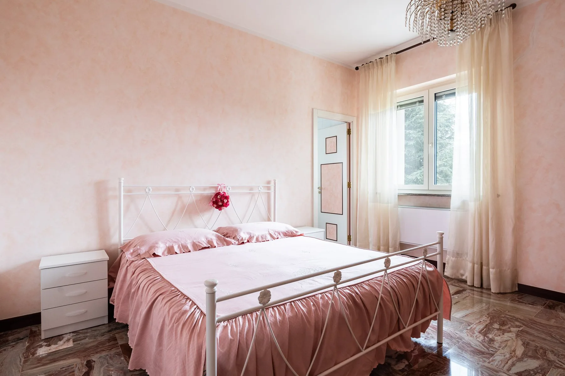 Un dormitorio con una cama de metal blanco vestida con ropa de cama rosa junto a una ventana con cortinas.