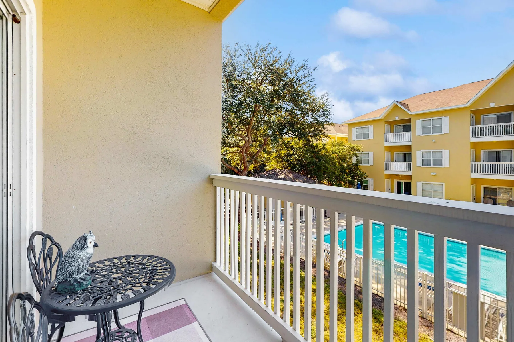 A balcony seating area overlooks a swimming pool and a yellow apartment building.