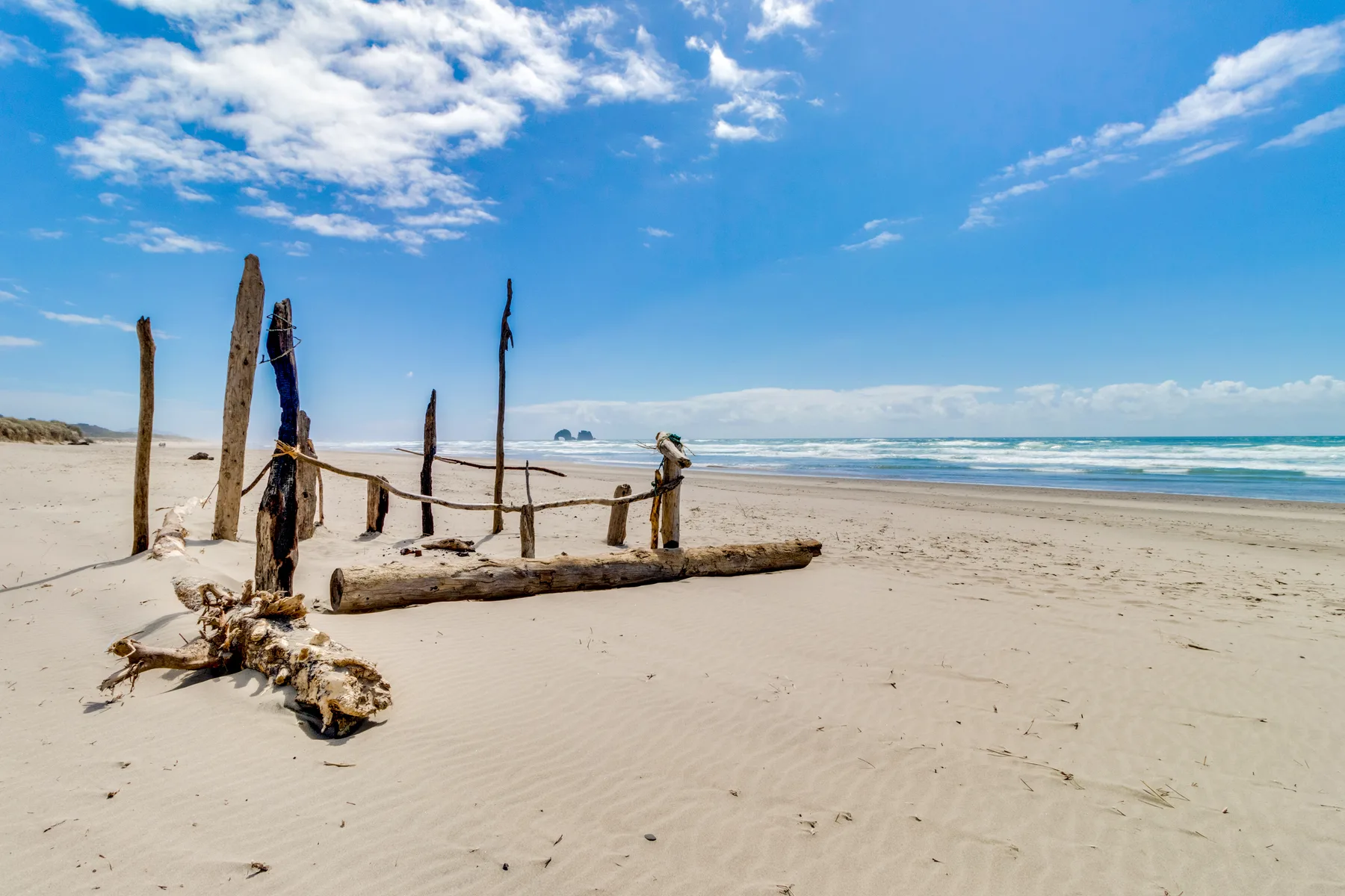 A sandy beach with driftwood posts and ocean waves under a partly cloudy sky.