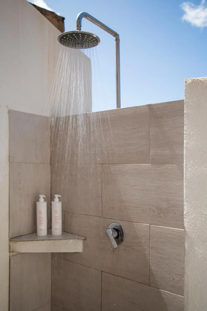 Una ducha al aire libre rocía agua contra una pared de azulejos bajo un cielo despejado.