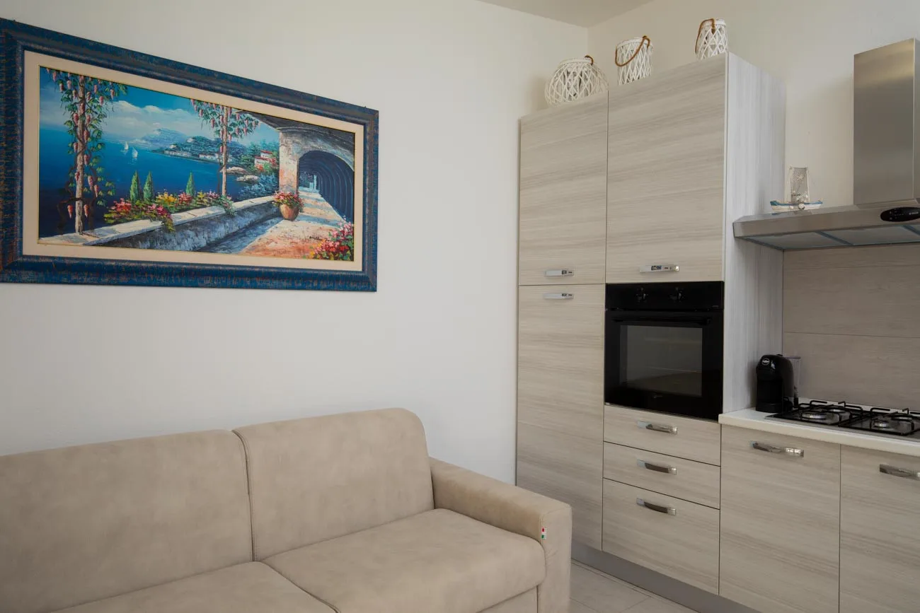Una zona de estar con asientos beige junto a una cocina compacta y un cuadro de paisaje.