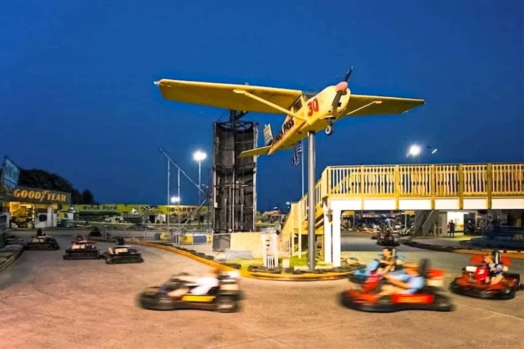 Una pista de karts al aire libre gira bajo un avión amarillo suspendido por la noche.