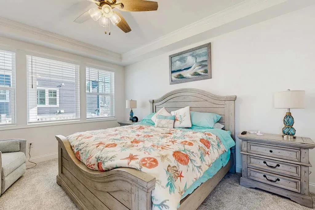 Un dormitorio luminoso con una cama cubierta con una colcha floral y ventanas grandes.
