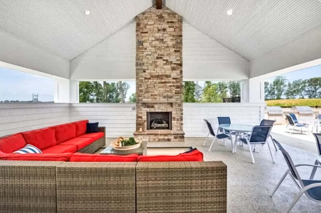 Un patio cubierto tiene asientos rojos frente a una chimenea central de piedra, con una mesa cercana.