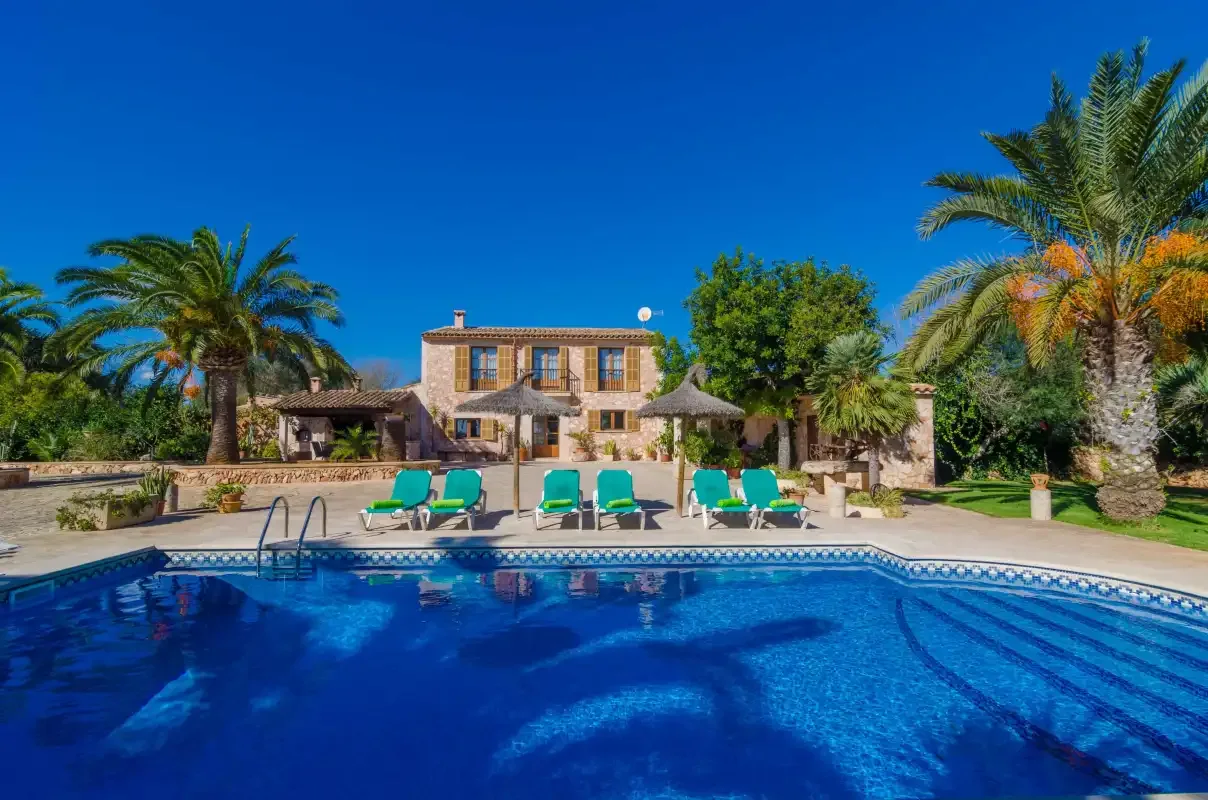 A swimming pool with lounge seating in front of a stone house and palm trees.