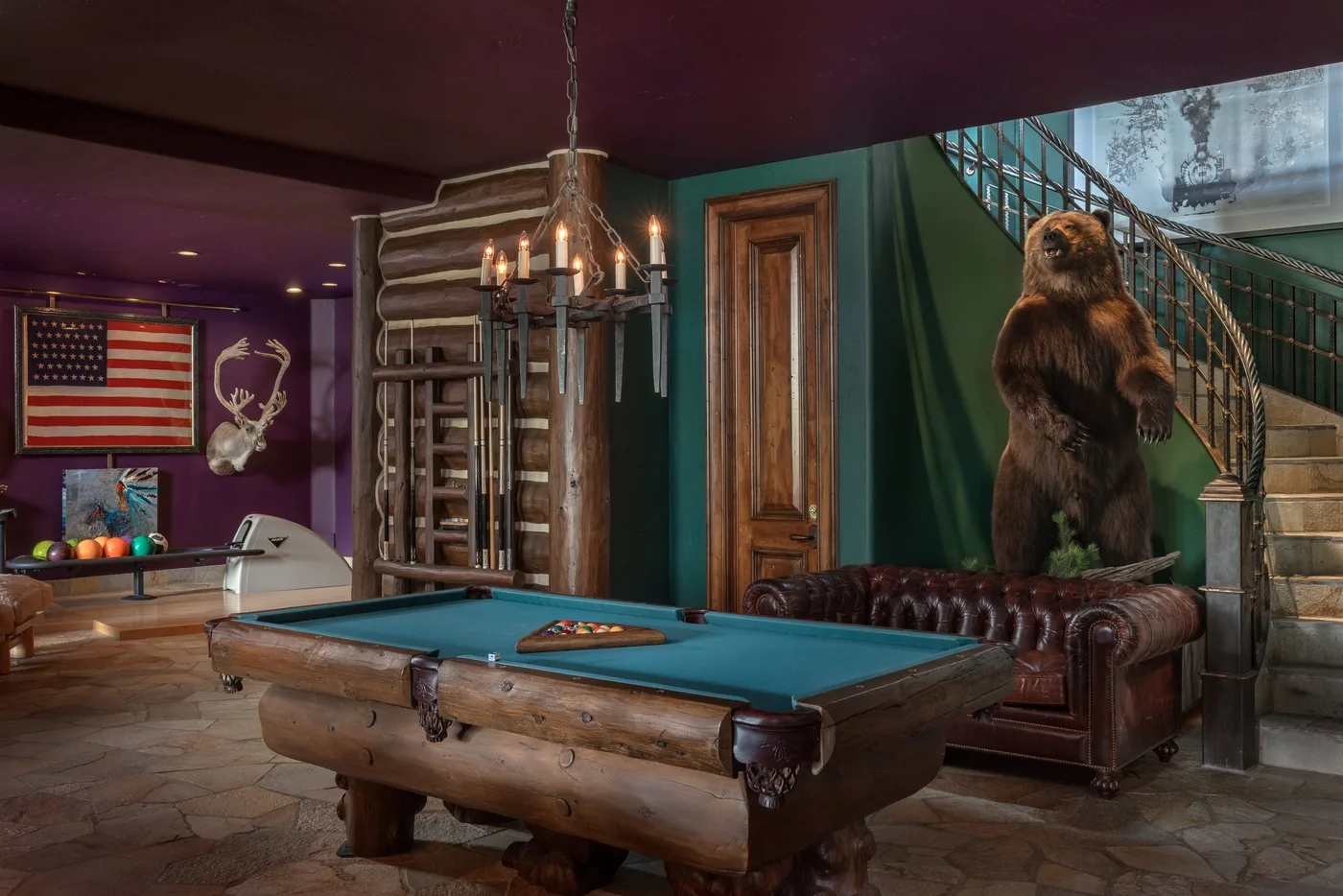 A rustic game room features a pool table and a large bear statue beside a staircase.