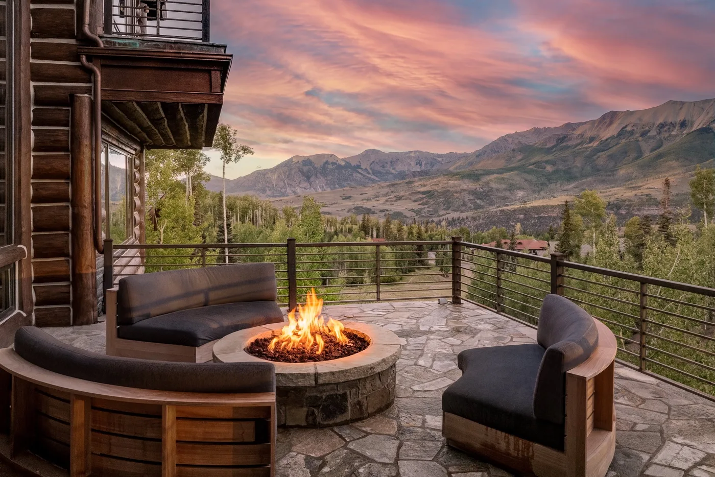 A stone patio with cushioned seating and a fire pit overlooks a mountain valley.