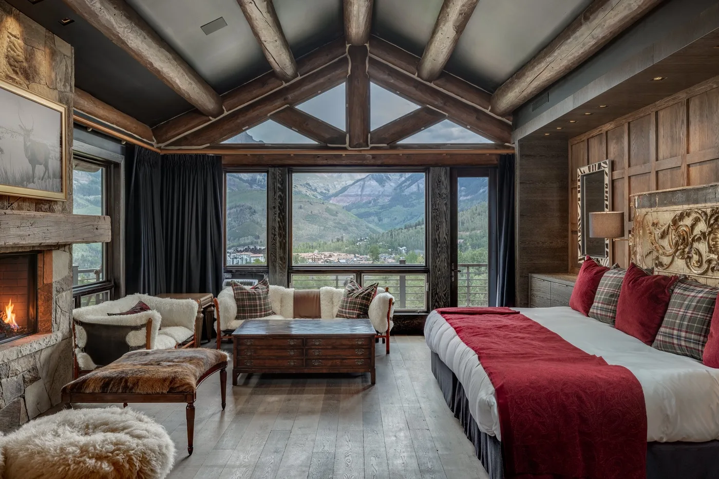 A bedroom with a bed, seating area, fireplace, and large windows overlooking mountains.