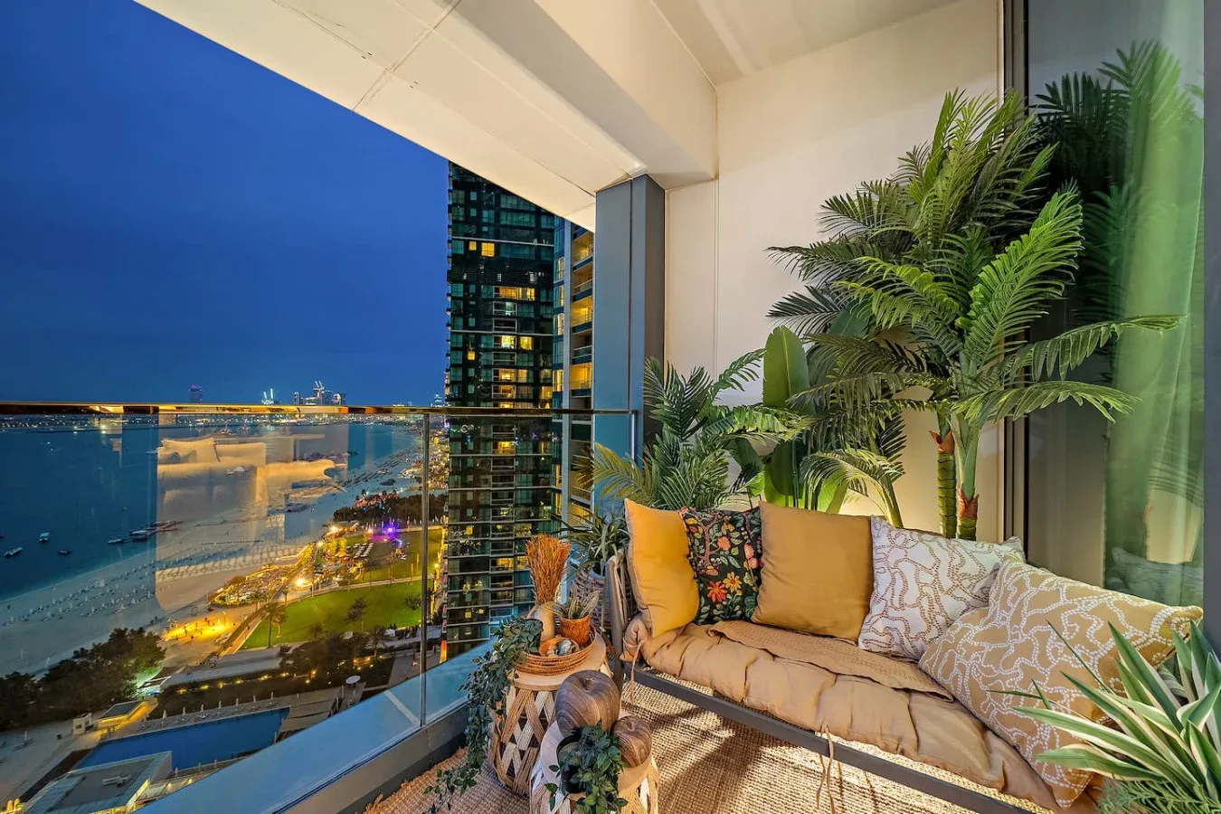 Una zona de estar en el balcón da al paseo marítimo al anochecer, con plantas junto a la barandilla de vidrio.