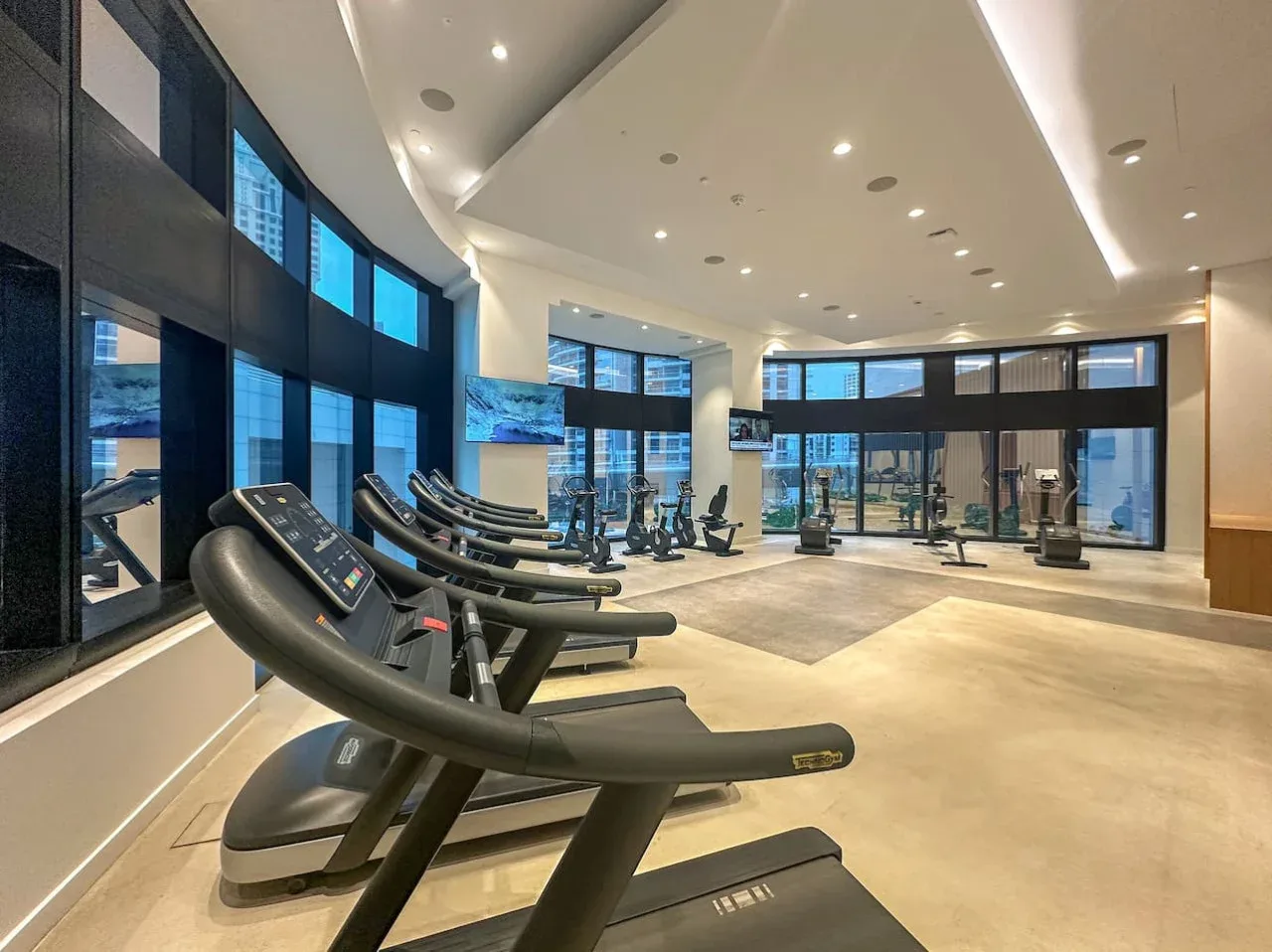 Un gimnasio amplio con cintas de correr frente a grandes ventanales.