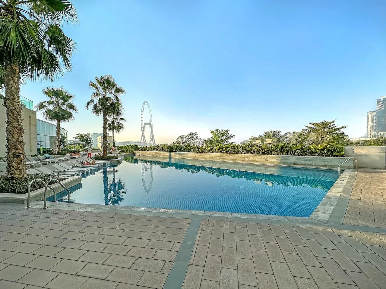Una piscina al aire libre está junto a una terraza pavimentada, con palmeras al fondo.