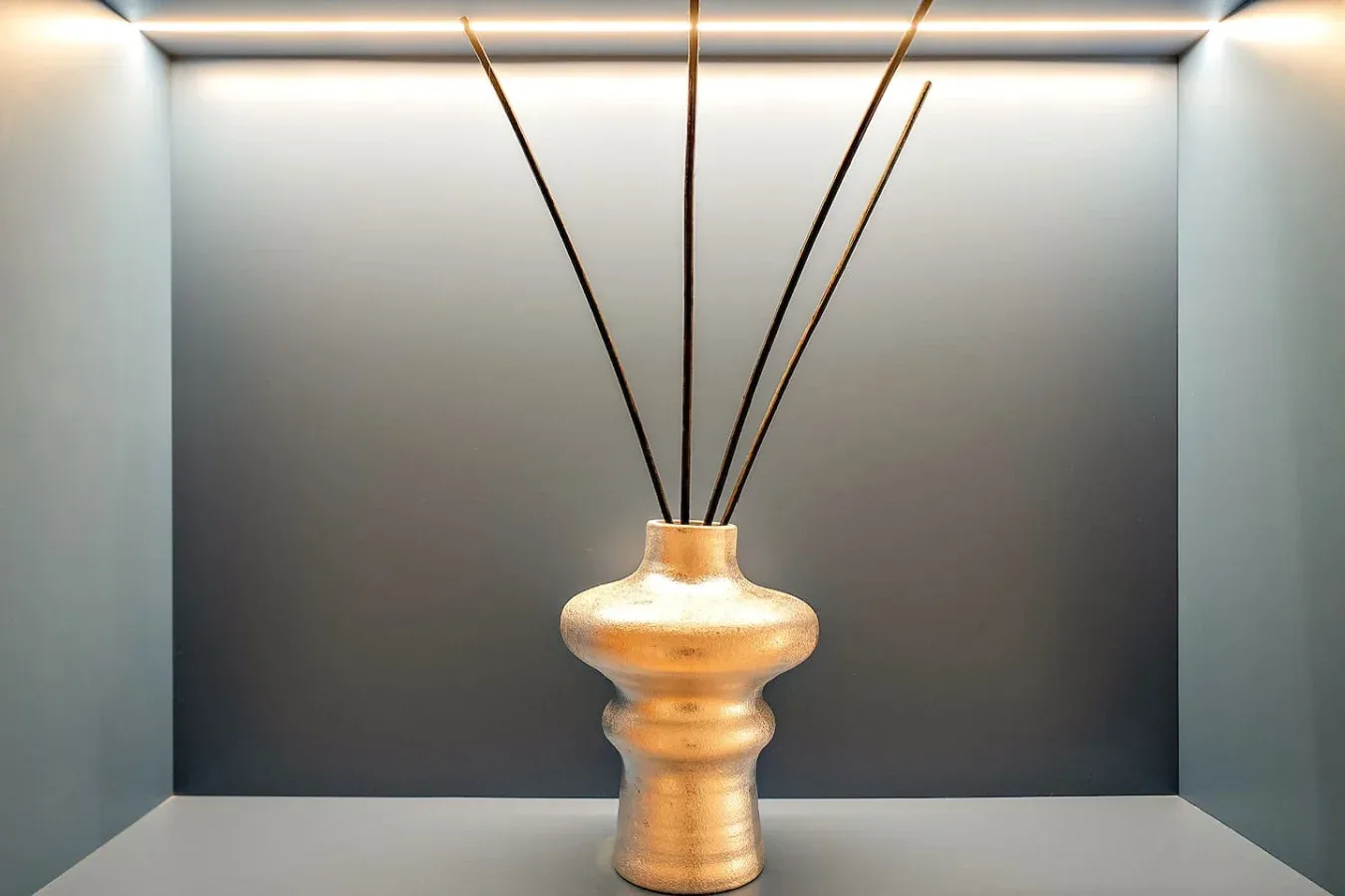 Un jarrón decorativo con tallos delgados está en un nicho de exhibición iluminado suavemente.
