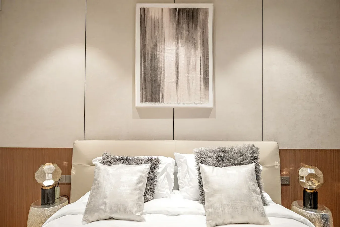 Una cama bien arreglada está bajo un cuadro abstracto, con lámparas a ambos lados.