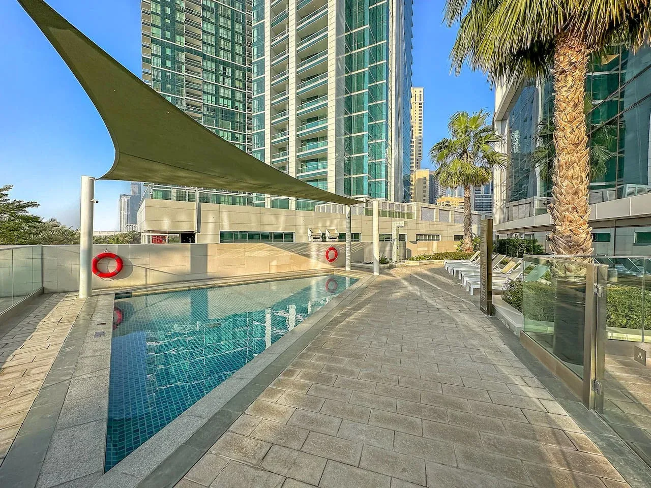 Una piscina al aire libre junto a una terraza pavimentada, bajo una lona y entre edificios altos.