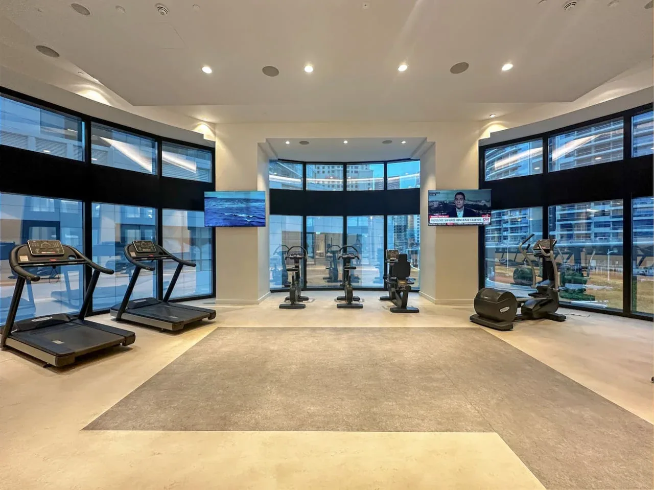 Un gimnasio con máquinas de ejercicio y grandes ventanales con edificios a la vista.