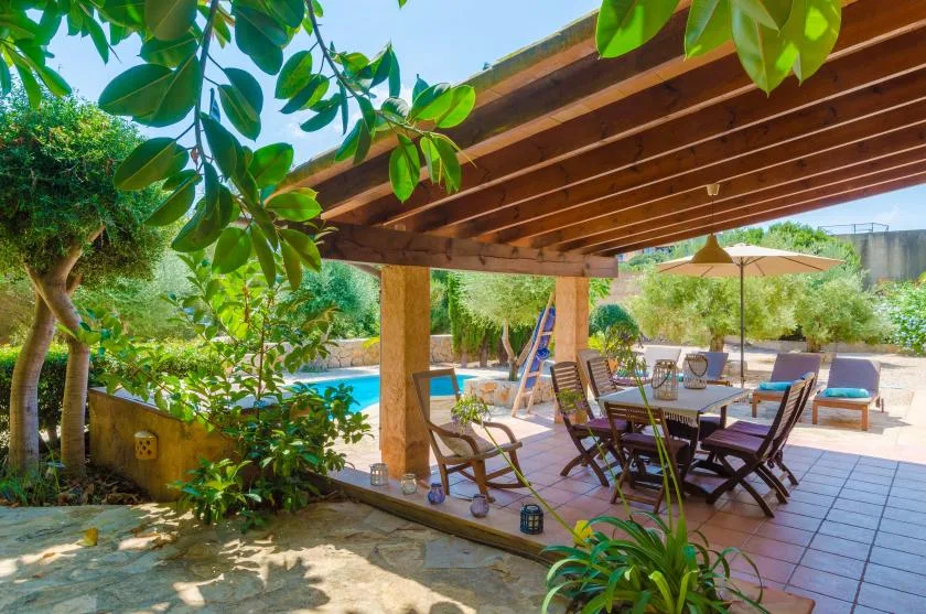 A shaded outdoor patio has a dining table and chairs beside a swimming pool.