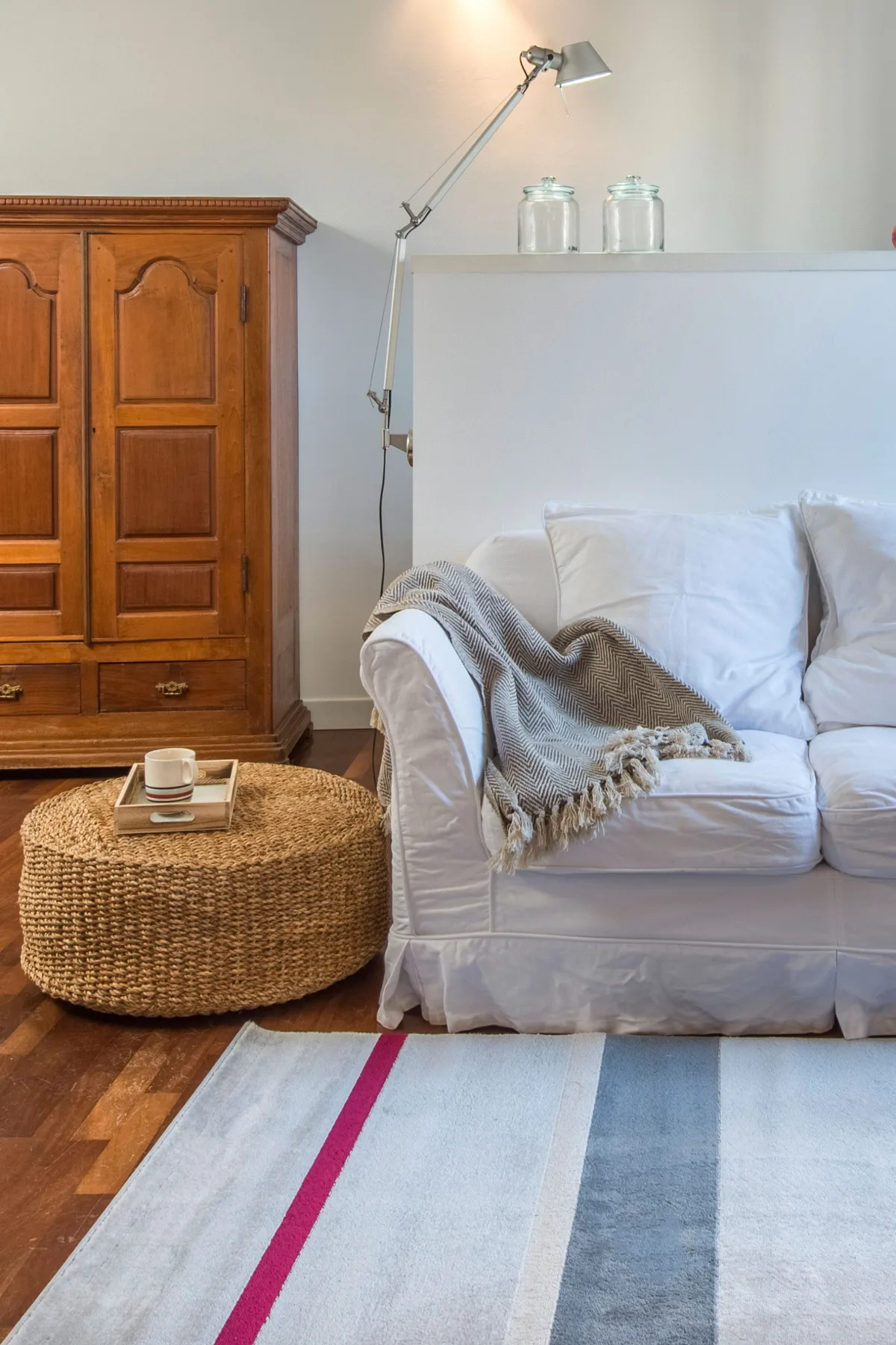 A living room with white seating, a woven ottoman, and a wooden cabinet.