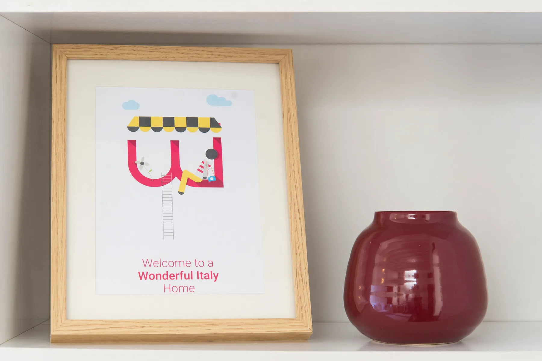A framed colorful print and a red glass vase sit on a white shelf.