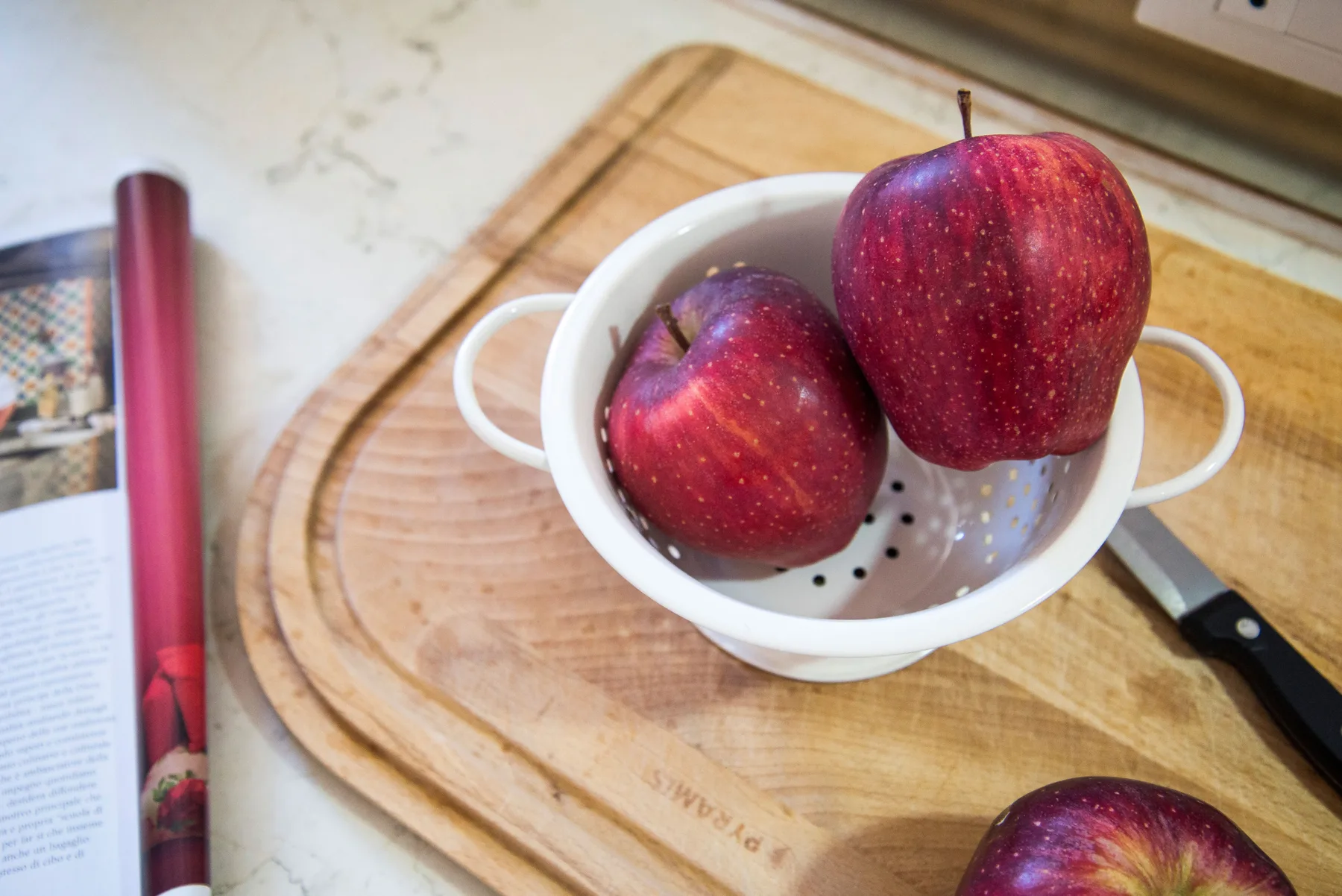 Apples sit in a white colander on a wooden cutting board.