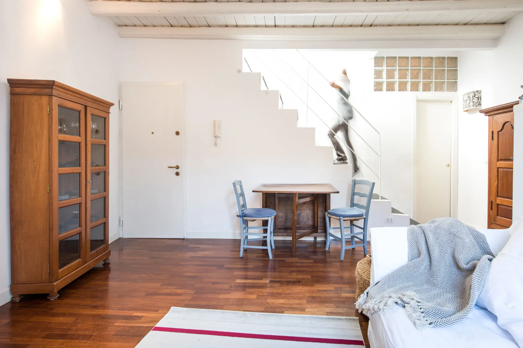 A bright living room with wooden furniture and a staircase along the back wall.