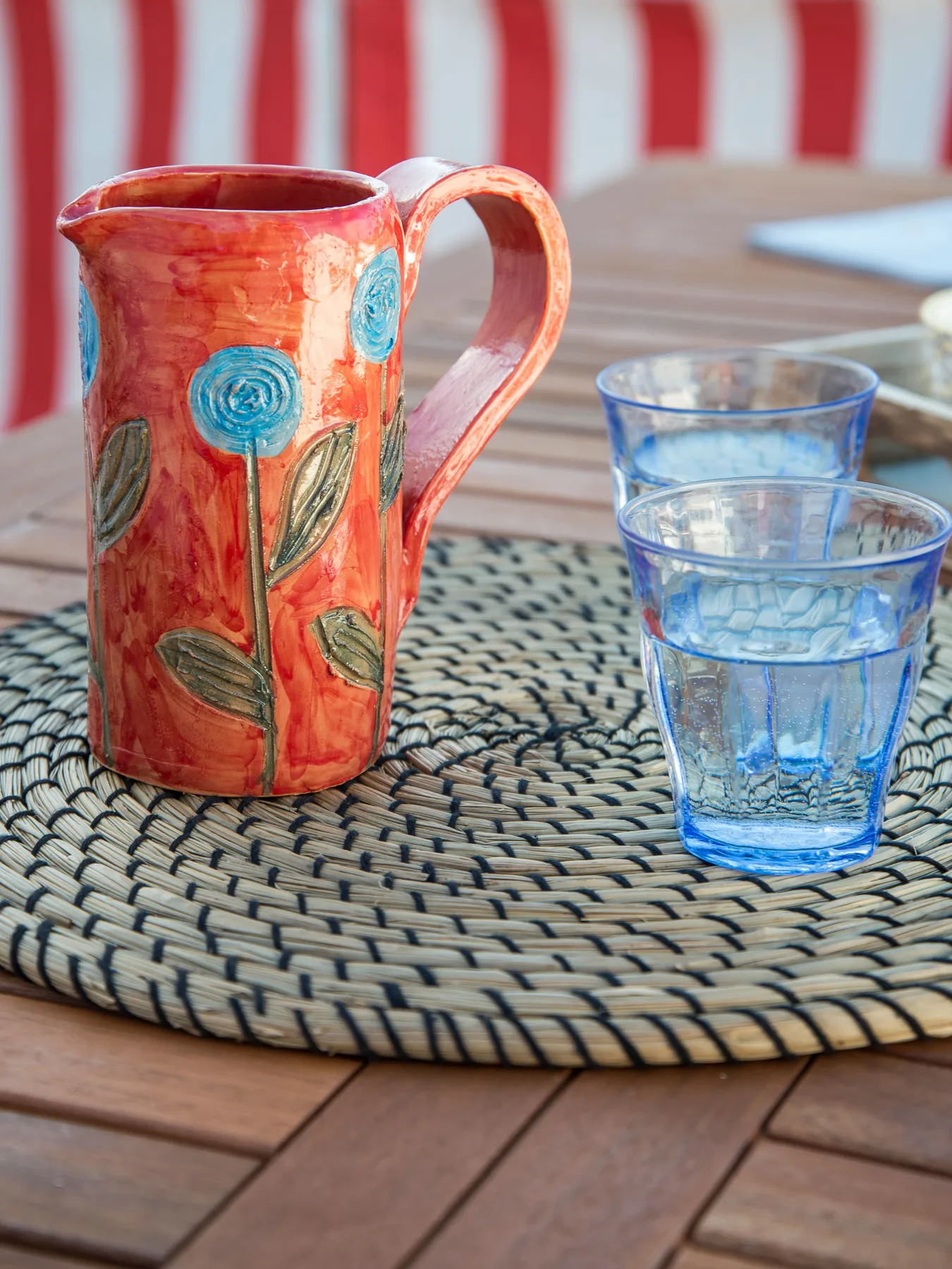 A colorful pitcher and blue drinking glasses sit on a woven mat atop a wooden table.