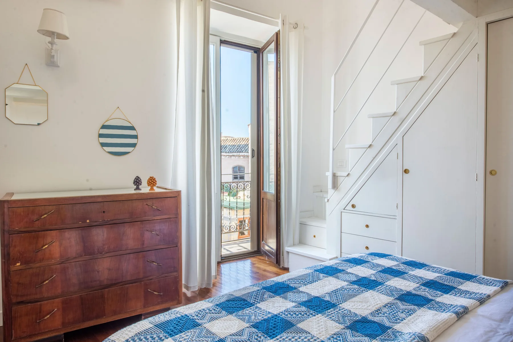 A bedroom with a blue patterned bedspread and a staircase beside the window.