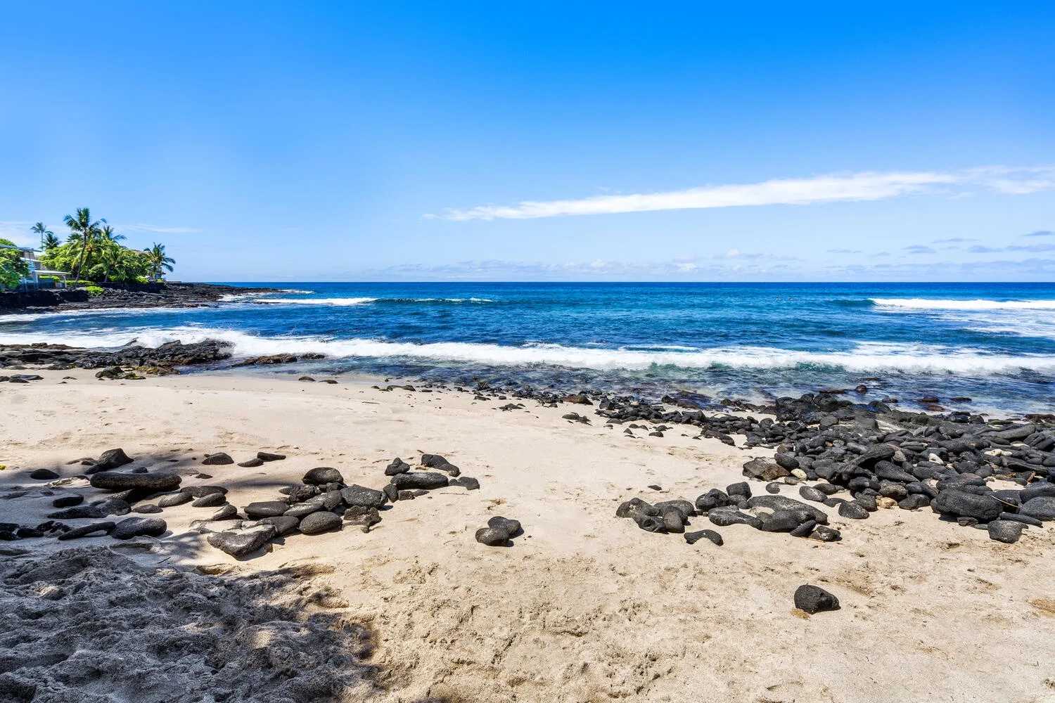 Una playa de arena con rocas oscuras se encuentra con olas azules bajo un cielo despejado.