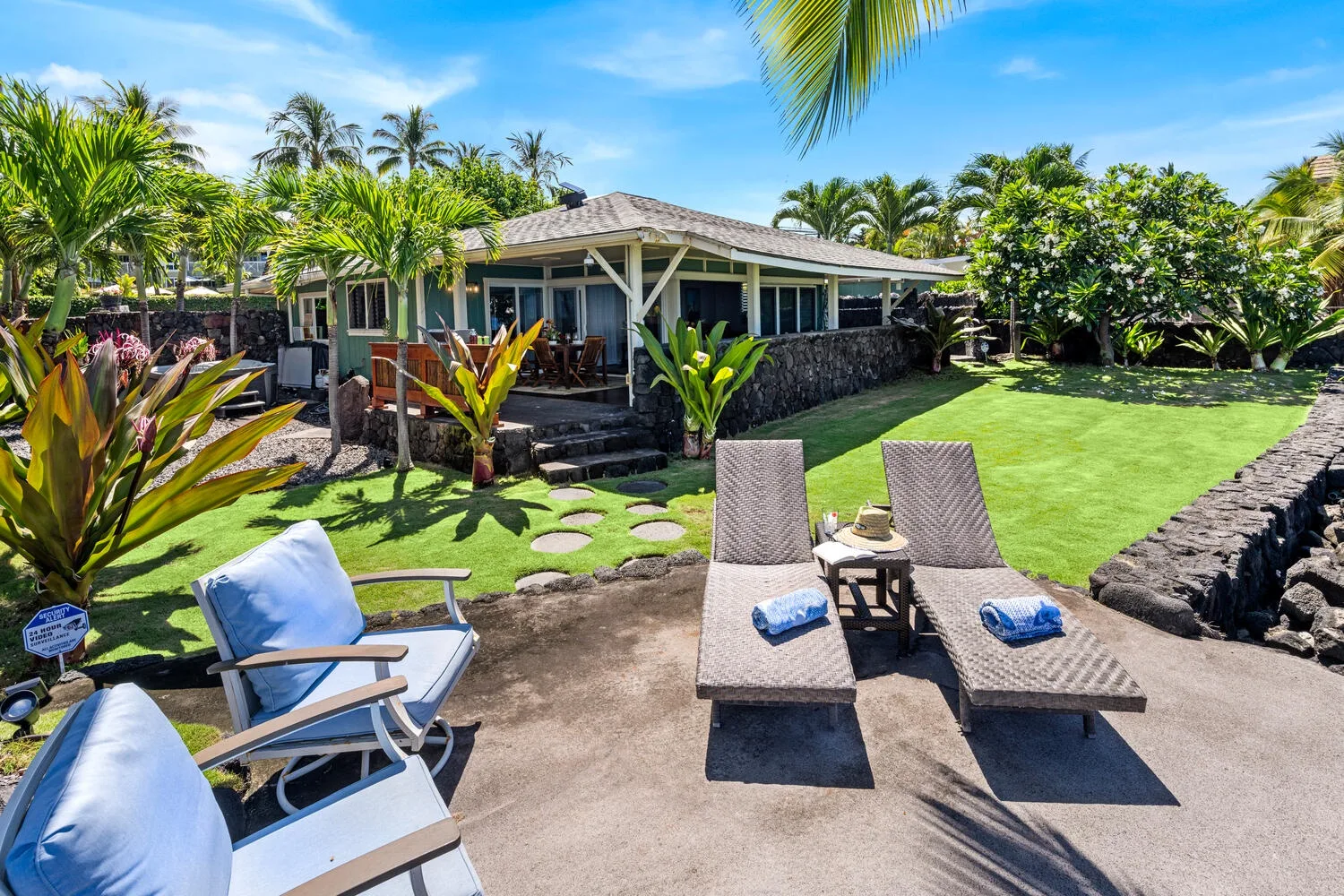 Un patio con jardín tropical tiene asientos junto a un césped y una casa baja.